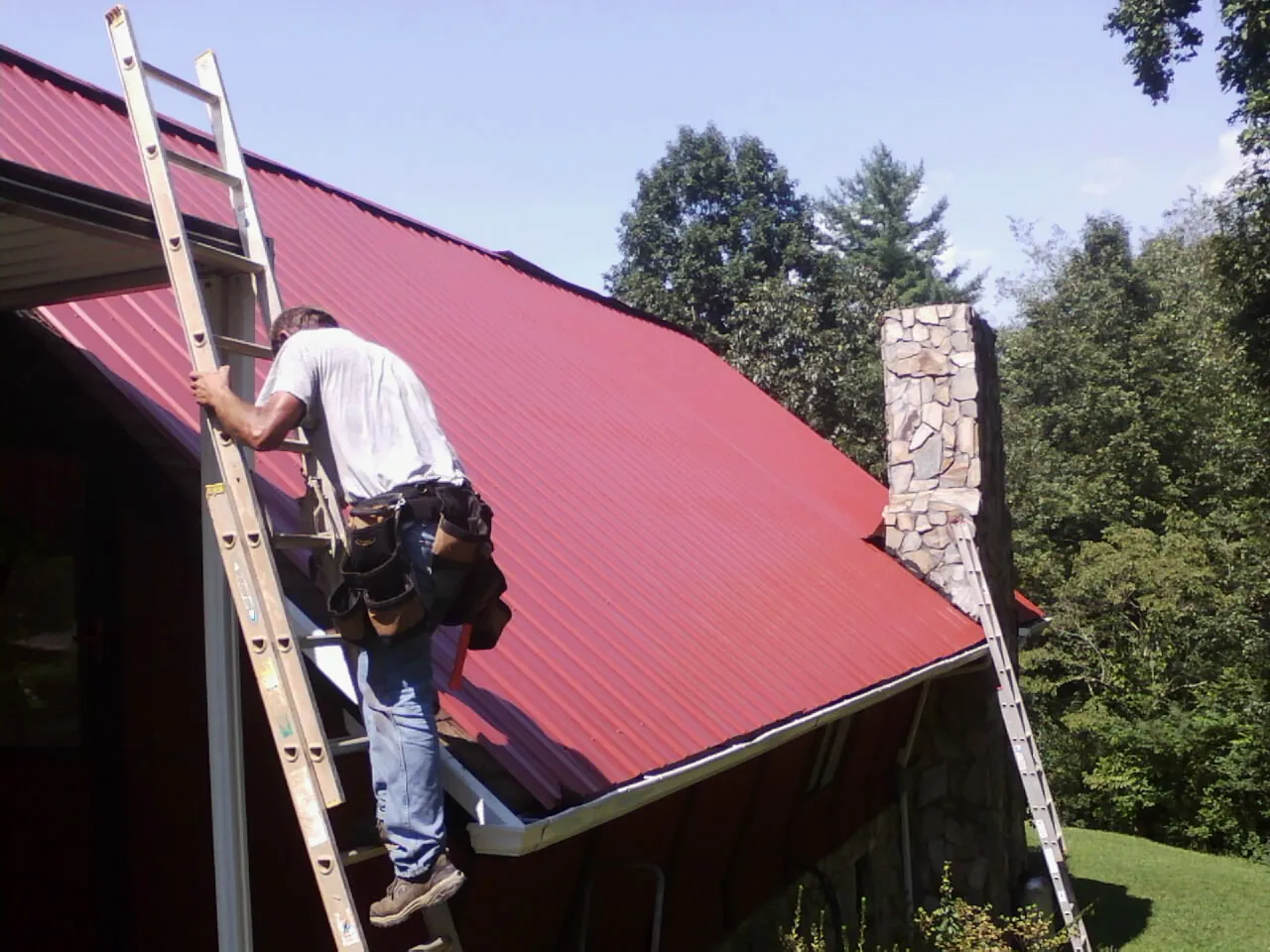 Roofer on a ladder beside a red metal roof and stone chimney — Soffit & Fascia Repair by Roofing Brothers in Flournoy